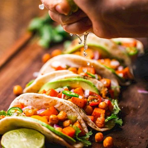 A hand squeezes lime juice over a row of tacos filled with chickpeas, avocado, and veggies, garnished with cilantro on a wooden board.