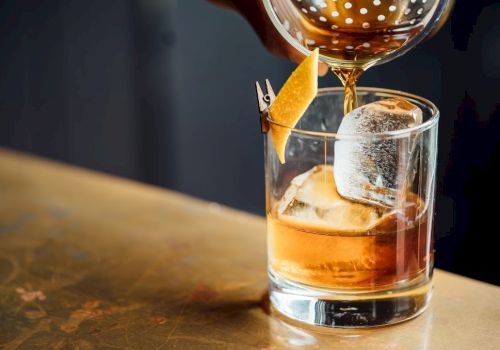 A cocktail being poured into a glass with a large ice cube and a lemon twist garnish.