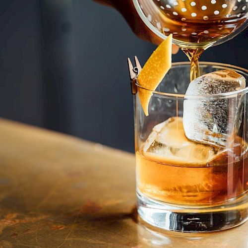 A cocktail being poured into a glass with a large ice cube and a lemon twist garnish.