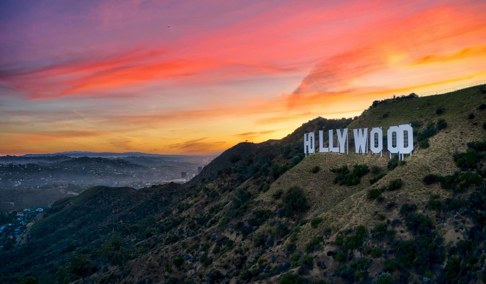 A famous hillside sign in Hollywood, Los Angeles, with a vibrant sunset sky in the background, overlooking the cityscape.