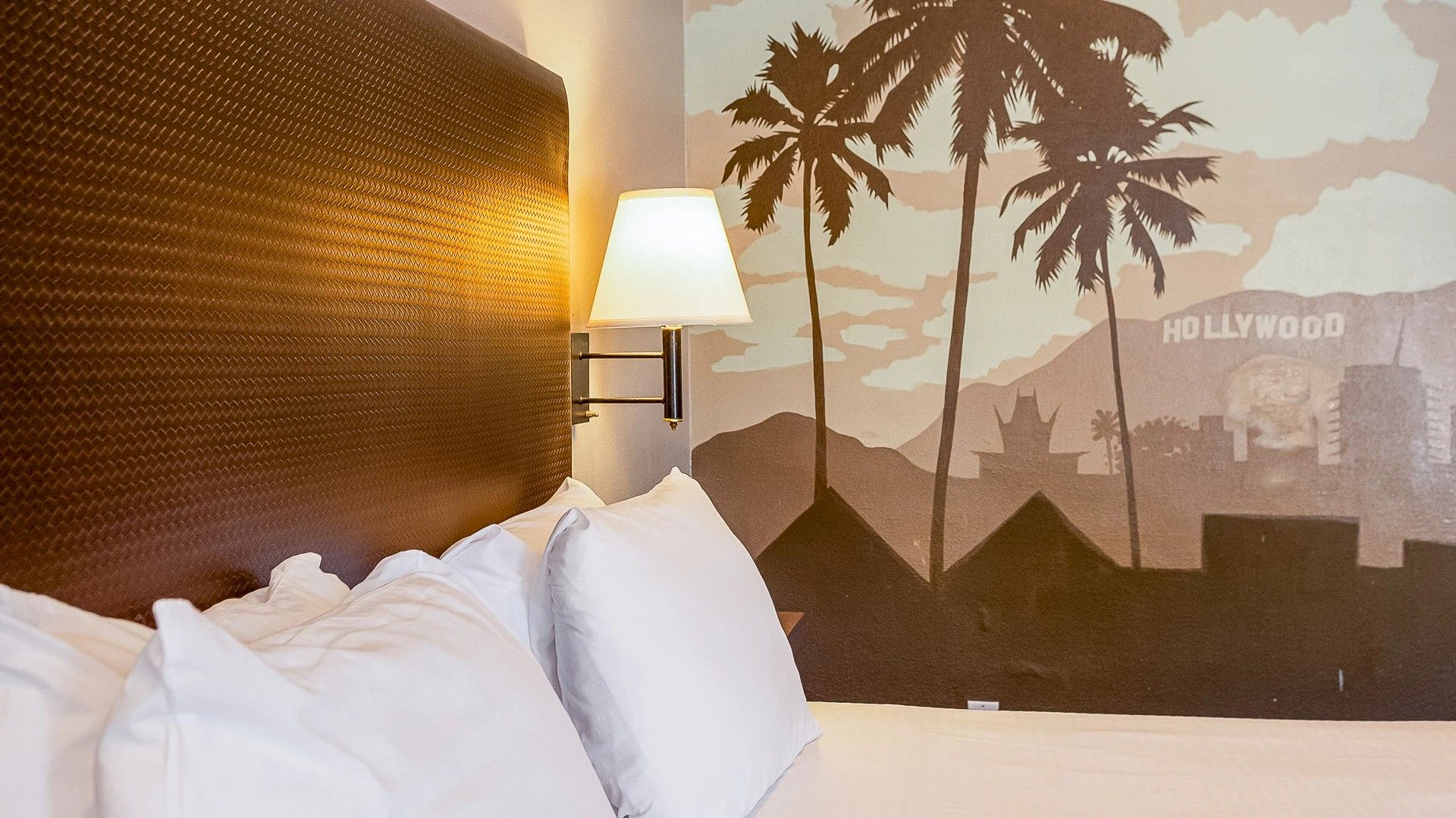 A hotel room with a headboard, pillows, wall lamp, and a mural featuring palm trees and the Hollywood sign.