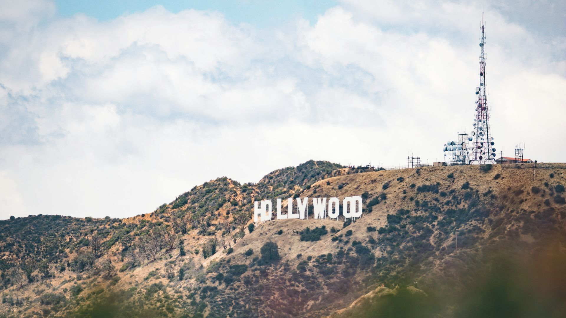 The image shows the Hollywood sign on a hillside with an antenna nearby under a blue, partly cloudy sky.