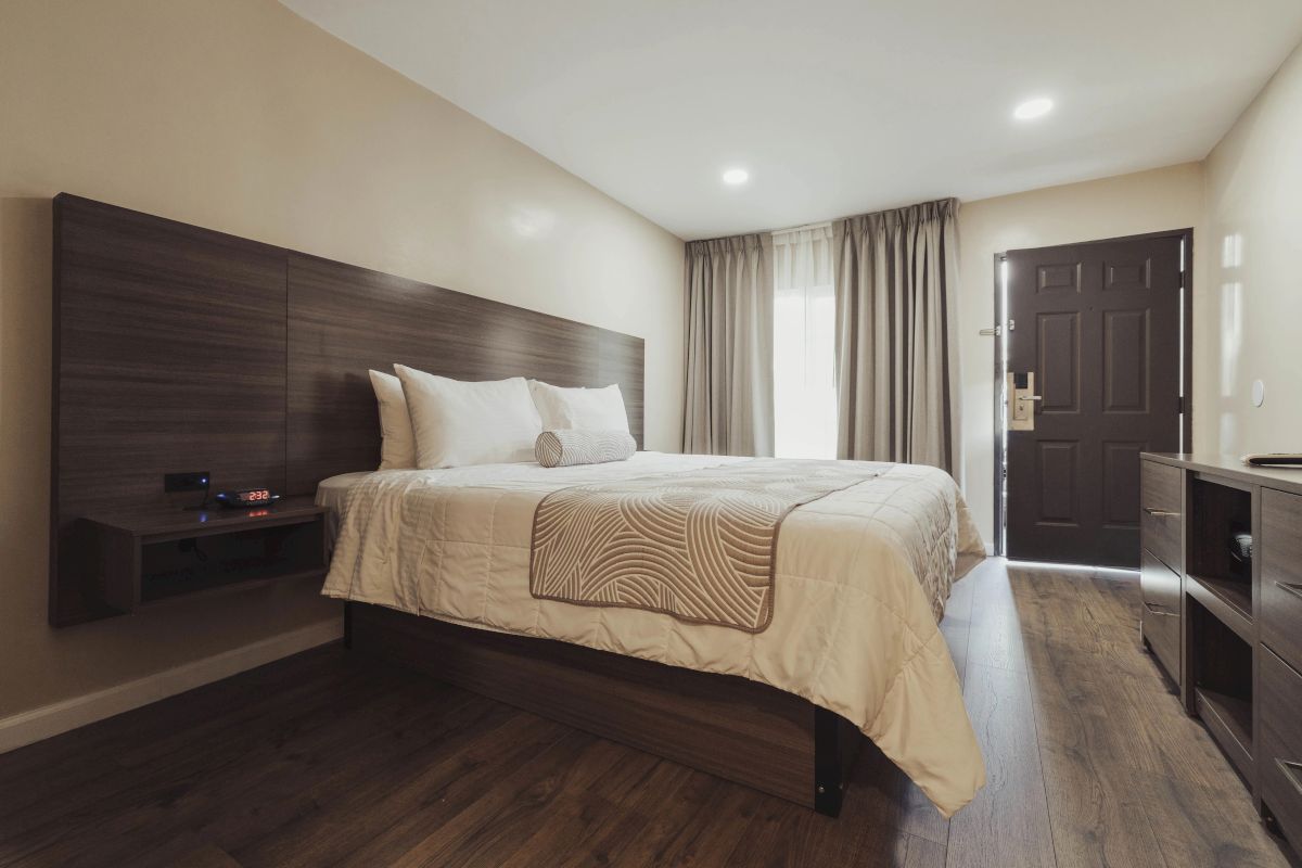 A hotel room with a large bed, nightstand, dresser, window, and door.