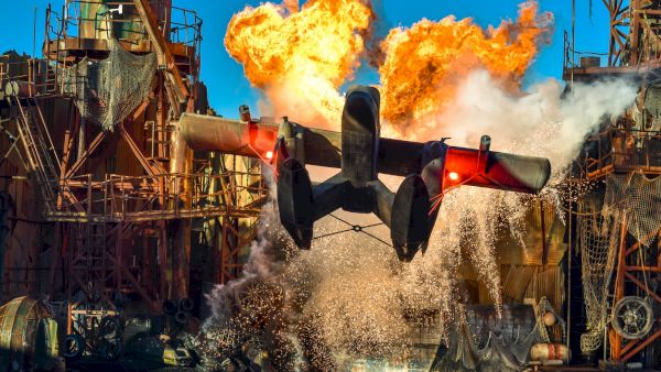 A seaplane flies over water with dramatic explosions and splashes in an industrial, action-packed scene.