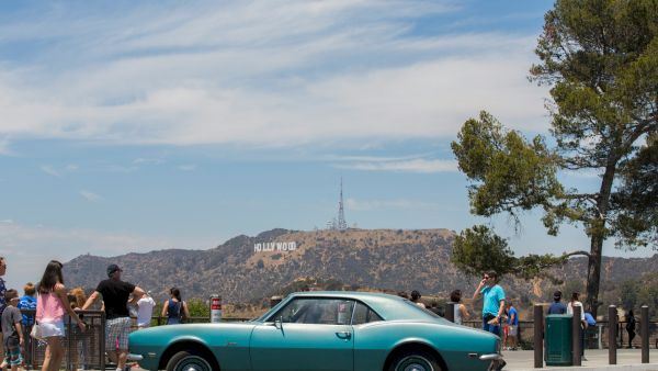 A vintage car is parked with people viewing the iconic Hollywood sign in the background, under a partly cloudy blue sky.