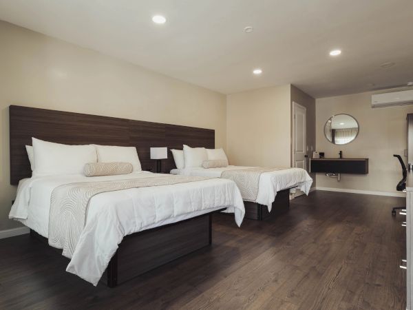 The image shows a hotel room with two double beds, modern decor, a wooden floor, and a wall-mounted mirror with a sink underneath.