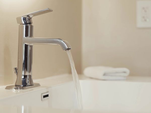 A modern chrome faucet with running water in a bathroom sink, with a folded towel in the background.
