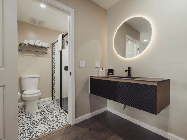 A modern bathroom with a round backlit mirror, black sink, patterned floor tiles, and a shower beside a toilet, with towels above.