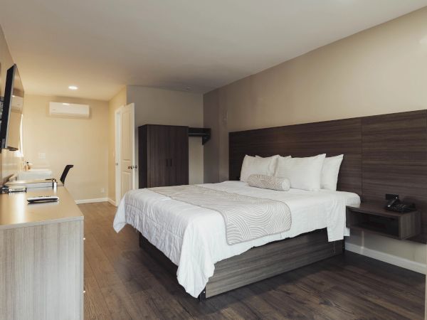 A modern hotel room with a neatly made bed, wall-mounted TV, desk, and chair. Cream walls and wooden floors add a cozy touch.