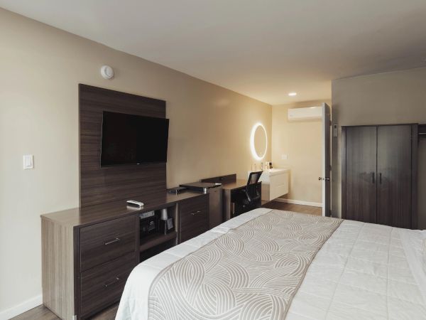 A neatly arranged bedroom features a bed, wall-mounted TV, dresser, desk with a glowing mirror, and a wardrobe in a soft-lit setting.