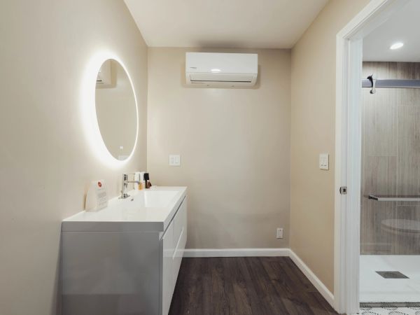 A bathroom with a round lighted mirror, counter with items, air conditioning unit, and doorway to a shower area with glass door.