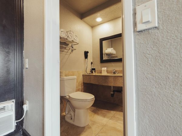 The image shows a bathroom with a toilet, sink, mirror, towels, and a hairdryer on the wall, viewed through an open doorway.