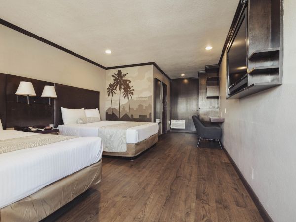 The image shows a hotel room with two beds, a large TV, a desk with a chair, a wood floor, and a palm tree mural on the wall.