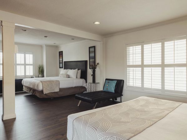 The image shows a spacious bedroom with two beds, a dark accent chair, and large windows with shutters, all in a contemporary style.