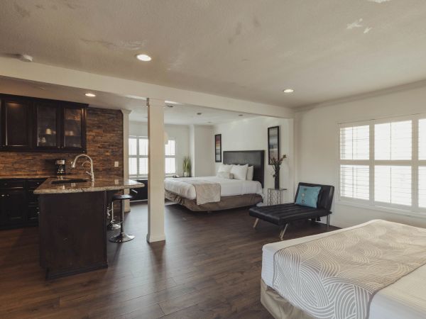 The image shows a spacious room with beds, a kitchenette, and seating area, featuring wooden flooring and bright lighting.