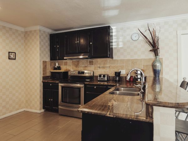 A modern kitchen with dark cabinetry, stainless steel appliances, a tiled backsplash, and a marble countertop with bar stools.