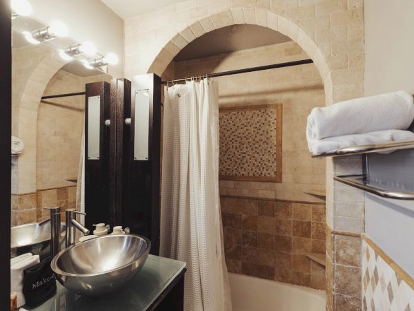 A bathroom featuring a vessel sink, tiled bathtub, shower curtain, mirrors, lighting, and neatly stacked towels on a shelf ends the scene.