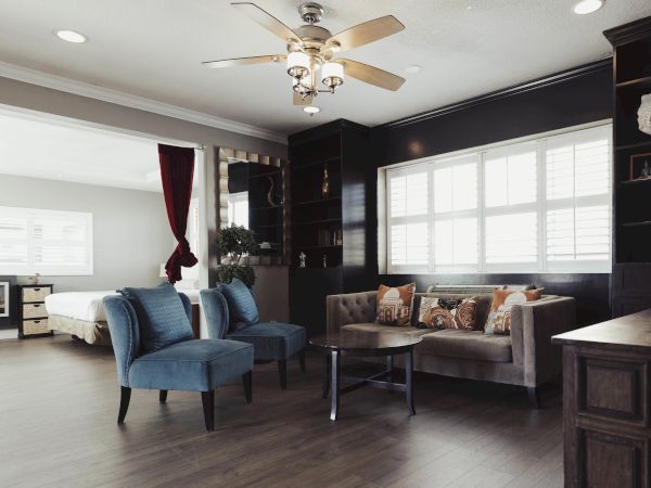 A modern living room with blue chairs, a beige sofa, wooden table, ceiling fan, and large window. Bedroom visible through an open doorway.