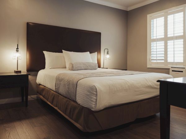 A neatly made bed in a cozy room with a wooden floor, side tables, lamps, and a window with shutters on the right side of the image.