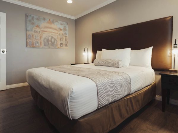 A cozy bedroom with a made bed, headboard, two wall lamps, and a painting of a building above the bed. The floor is wooden.