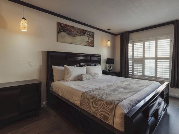 A cozy bedroom with a large bed, elegant lighting, a painting above the headboard, and a window with shutters and curtains.