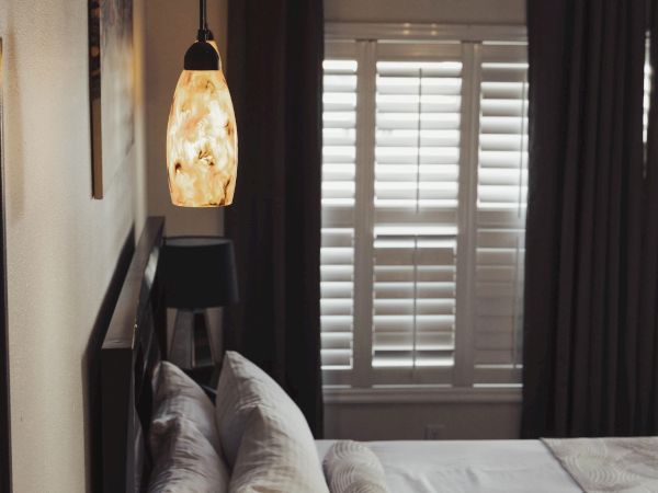 A cozy bedroom with a bed, pillows, a pendant lamp, and a window with shutters and curtains.
