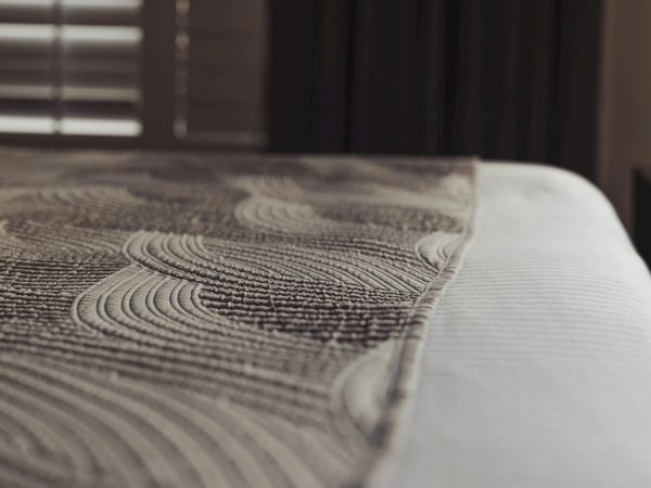 A neatly made bed with a textured, patterned bedspread. The room has soft lighting and window blinds on the left side.