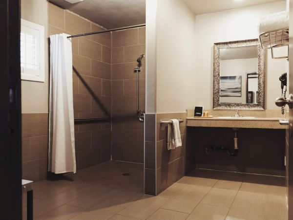 The image shows a modern, accessible bathroom with a roll-in shower, curtain, sink, mirror, and towel racks on beige tiled walls.