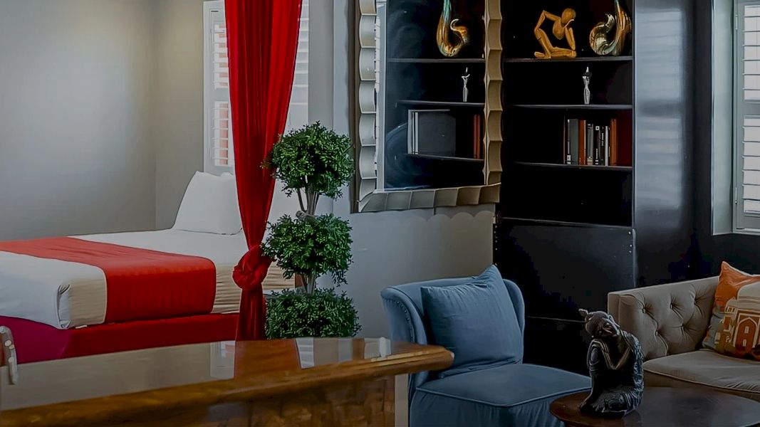 A modern living space with a bed, red curtains, seating area, and shelves. Cozy decor and natural light enhance the ambience.