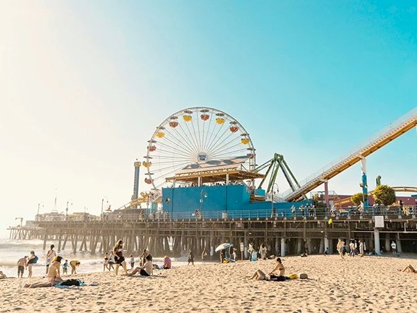 The image shows a beach scene with people relaxing and playing. A ferris wheel and roller coaster are on a pier under a clear blue sky.