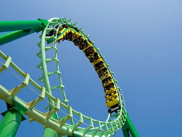A roller coaster with green supports and a yellow train is looping against a clear blue sky, full of excited riders.