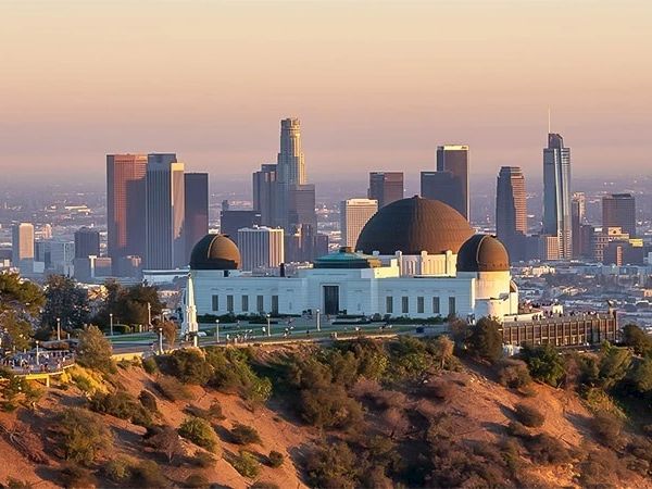 The image shows Griffith Observatory on a hillside with the Los Angeles skyline in the background during sunset.