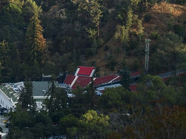 The image shows an outdoor amphitheater with red seats surrounded by trees in a hilly area.