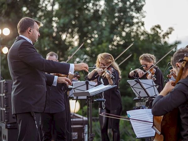 An outdoor orchestra performance with a conductor and string musicians playing instruments, set against a backdrop of trees and stage lighting.