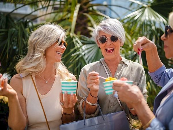 Three people are enjoying ice cream outdoors, smiling and chatting, surrounded by greenery and sunlight.