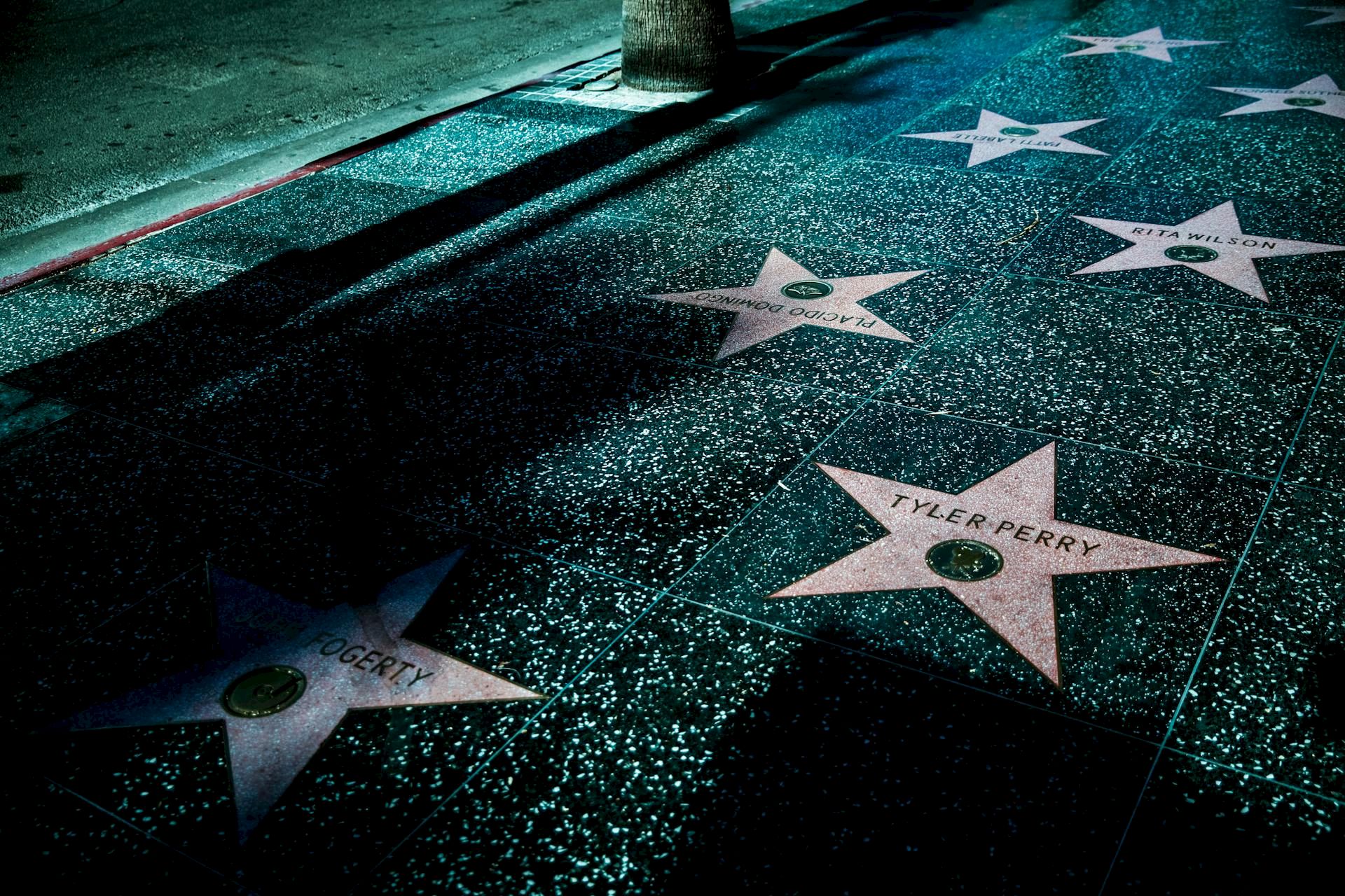The image shows stars on the Hollywood Walk of Fame, with names engraved on them, set against a dark, shadowy sidewalk.
