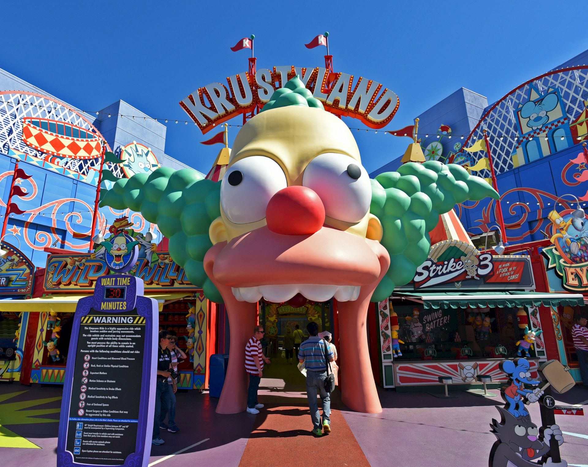 A large entrance resembling a clown's face with "Krustyland" sign, set in an amusement park with vibrant colors and designs.