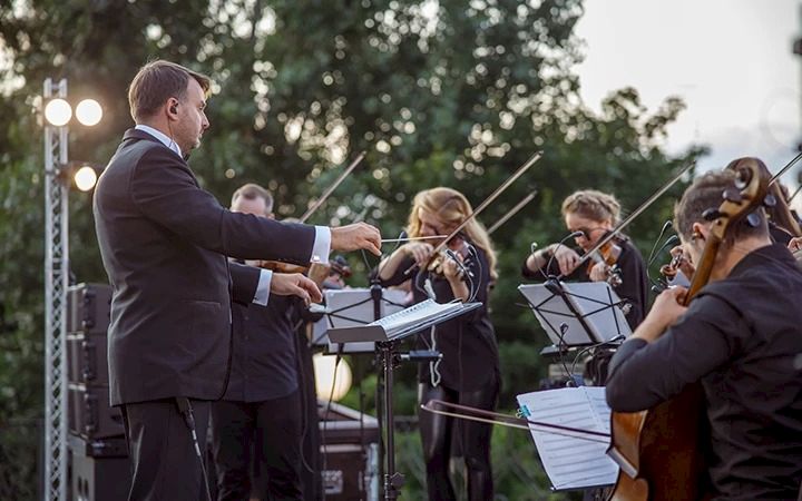 An orchestra performs outdoors with a conductor leading musicians playing string instruments.