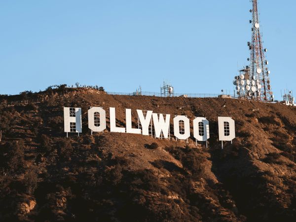The image shows the iconic Hollywood sign on a hillside with a large radio tower nearby, set against a clear blue sky.