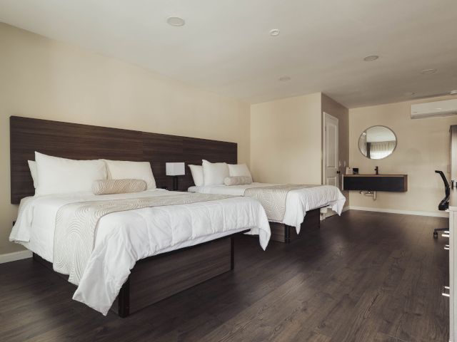 A wide view of a hotel room with two beds, a dark wood floor, and light-colored walls.
