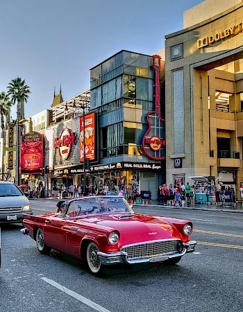 A classic red convertible drives past the Hard Rock Cafe and Dolby Theatre in a bustling street scene.