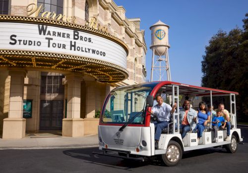 A tram with people is in front of the Warner Bros. Studio Tour Hollywood entrance and a water tower with a WB logo in the background.