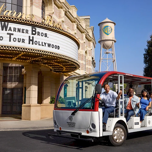 A tram with people is in front of the Warner Bros. Studio Tour Hollywood entrance and a water tower with a WB logo in the background.
