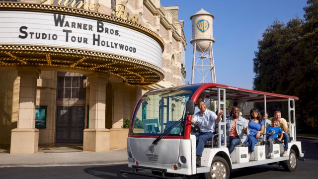 A tram with people is in front of the Warner Bros. Studio Tour Hollywood entrance and a water tower with a WB logo in the background.