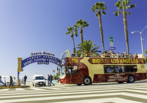 A Big Bus Los Angeles tour bus at Santa Monica Pier entrance, with palm trees and people around, under a clear blue sky.