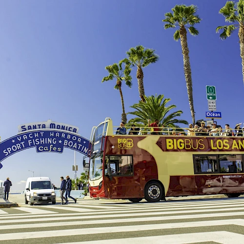 A Big Bus Los Angeles tour bus at Santa Monica Pier entrance, with palm trees and people around, under a clear blue sky.