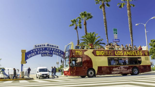A Big Bus Los Angeles tour bus at Santa Monica Pier entrance, with palm trees and people around, under a clear blue sky.