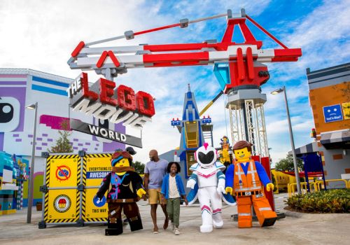 People with LEGO character mascots pose at "The LEGO Movie World" entrance in a theme park setting.