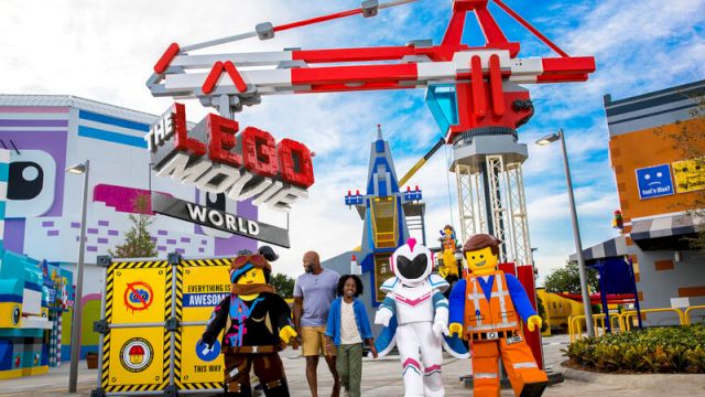 People with LEGO character mascots pose at "The LEGO Movie World" entrance in a theme park setting.