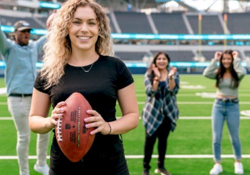 A person holding a football on a field, with three individuals cheering in the background inside a stadium.